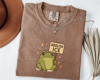 May include: A brown short-sleeved t-shirt featuring a graphic of a green frog holding a sign that reads ABOLISH ICE, surrounded by small colorful stars. The shirt has a crew neckline and a Comfort Colors brand label printed on the inside collar.