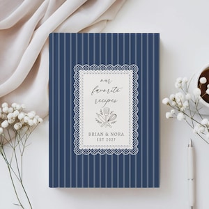 May include: A navy blue recipe book with white vertical pinstripes. The centre features a white rectangular label with a decorative lace border, the text "our favourite recipes", a graphic of a spatula, rolling pin, and whisk, and "BRIAN & NORA EST 2027".