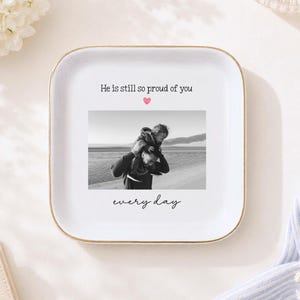 May include: A square white ceramic trinket dish with a gold-toned rim. The center features a black and white photograph of a person carrying a child on their shoulders, with the text "He is still so proud of you" above and "every day" below.