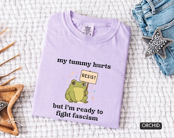May include: A light purple t-shirt with the text "my tummy hurts but I'm ready to fight fascism." A cartoon frog holds a sign that says "RESIST." The shirt is next to a pair of blue jeans and a star-shaped accessory.