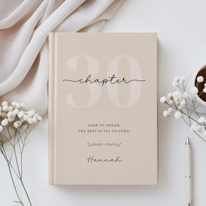 Puede incluir: Un libro de tapa dura beige con el texto grande 30, la palabra chapter en letra cursiva y el texto DARE TO DREAM. THE BEST IS YET TO COME. seguido de un pequeño adorno floral y el nombre Hannah en letra cursiva en la portada.