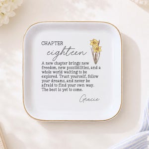 May include: A square white ceramic trinket dish with a gold-toned rim. The center features a yellow daffodil illustration and black text reading: CHAPTER eighteen, A new chapter brings new freedom, new possibilities, and a whole world waiting to be explored. Trust yourself, follow your dreams, and never be afraid to find your own way. The best is yet to come. Gracie.