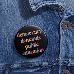 May include: A black button with the text "democracy demands public education" in colorful letters. The button is pinned to a blue denim jacket.