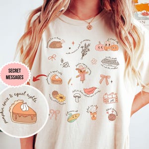 May include: A cream-coloured t-shirt featuring autumn-themed illustrations and text. The design includes pumpkins, pie, a scarecrow, and the words "pumpkin spice & equal rights." The shirt has a relaxed fit.