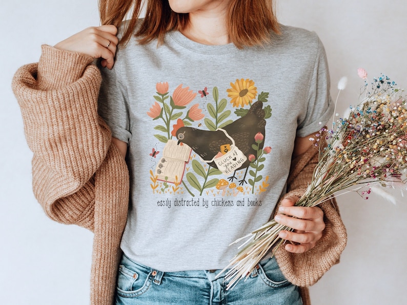 May include: A gray t-shirt with a black chicken holding a book. The chicken has a sign that says "Clutch Your Book & Read". The shirt also has a floral design and the text "easily distracted by chickens and books".