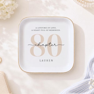 May include: A square white ceramic trinket dish with rounded corners and a gold-toned rim. The centre features the text A LIFETIME OF LOVE. A HEART FULL OF MEMORIES. above the large beige number 80, the word chapter in script, and LAUREN.