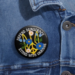 Puede incluir: Un botón negro con un signo de paz amarillo rodeado de flores. El texto en el botón dice "Defender la democracia Apoyar a Ucrania".