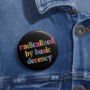 Op de afbeelding: Een ronde zwarte pin-back button met de tekst "radicalized by basic decency" in een kleurrijk, gestileerd lettertype met letters in tinten roze, geel en lichtblauw, bevestigd op een blauwe spijkerstof met zichtbare stiksels en knopen.