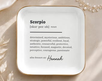 May include: A square white ceramic trinket dish with rounded corners and a gold-toned rim. The center features black text defining Scorpio as determined, mysterious, ambitious, strategic, powerful, resilient, loyal, authentic, and more, ending with also known as: Hannah.