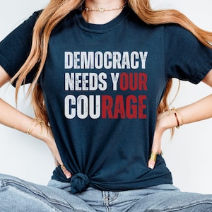 May include: Navy blue t-shirt with the text "DEMOCRACY NEEDS YOUR COURAGE" in white and red block letters. The shirt is knotted at the waist and worn by a person with long, light brown hair.