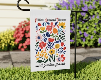 May include: A white garden flag hanging on a black metal stand. The flag features a colorful floral pattern in red, yellow, blue, and pink. The text at the top reads "freedom democracy tolerance" and the text at the bottom reads "and justice for all".