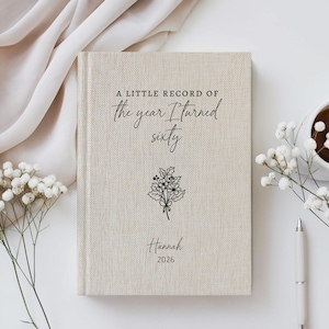 Può includere: Un diario con copertina rigida in tessuto color beige, caratterizzato dalla scritta nera A LITTLE RECORD OF the year I Turned sixty, una piccola illustrazione floreale a linee, il nome Hannah e l'anno 2026 sulla copertina frontale.