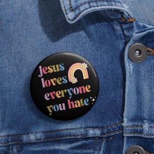 Könnte beinhalten: Ein schwarzer Button mit dem Text "Jesus loves everyone you hate" in bunten Buchstaben. Über dem Text befindet sich eine Regenbogen- und Gänseblümchen-Grafik. Der Button ist an einer Jeansjacke befestigt.