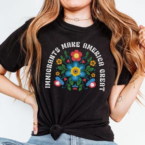 May include: Black t-shirt with the text "IMMIGRANTS MAKE AMERICA GREAT" in a white arch. The shirt features a colorful floral design in the center, with blue, yellow, red, and green accents. The shirt is tied at the waist.