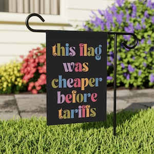 May include: A black garden flag with the text "this flag was cheaper before tariffs" in colorful, stylized letters.