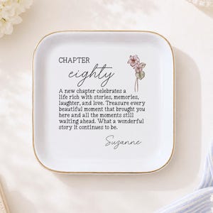 Puede incluir: Un plato cuadrado de cerámica blanca con esquinas redondeadas y un borde dorado. El centro presenta una ilustración floral y el texto: CHAPTER eighty, A new chapter celebrates a life rich with stories, memories, laughter, and love.