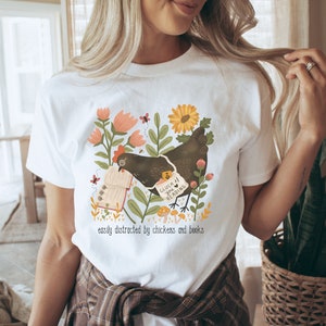 May include: White t-shirt with a graphic of a chicken reading a book. The graphic includes flowers, butterflies, and mushrooms. The text on the shirt reads "Good luck if you're reading" and "easily distracted by chickens and books".