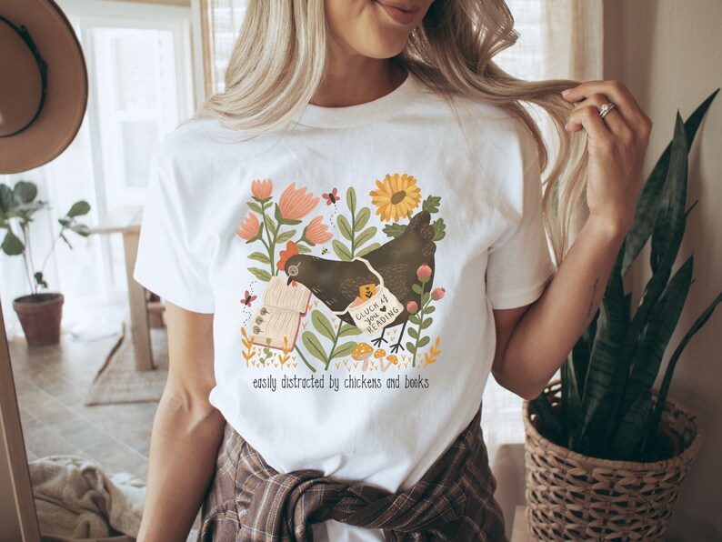 May include: White t-shirt with a graphic of a chicken reading a book. The graphic includes flowers, butterflies, and mushrooms. The text on the shirt reads "Good luck if you're reading" and "easily distracted by chickens and books".