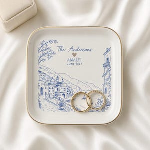 Puede incluir: Un plato cuadrado de cerámica blanca con borde dorado, que presenta una ilustración en tinta azul de una ciudad costera de Amalfi. El texto dice The Andersons, un icono de corazón, AMALFI y JUNE 2027. Dos anillos descansan sobre la superficie.