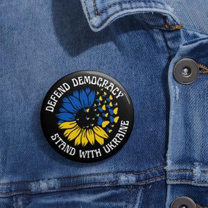 May include: A black button featuring a sunflower design. The sunflower is yellow and black with blue butterflies flying around it. The text on the button reads "Defend Democracy Stand With Ukraine".