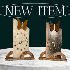 May include: Two wooden clocks with butterfly accents. The clocks have Roman numeral faces and are on a white marble surface.