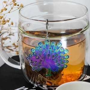 May include: A glass tea infuser with a peacock-shaped metal tea strainer. The peacock is iridescent with shades of blue, green, and purple. The infuser is hanging from a chain and is in a glass mug filled with tea.