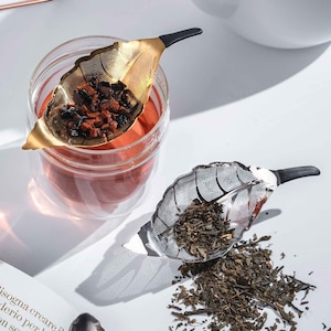 May include: Two gold and silver leaf-shaped tea infusers. The gold infuser is filled with loose leaf tea and the silver infuser is filled with loose green tea. The tea infusers are on a white surface.