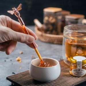 May include: A copper-coloured honey dipper with a bee-shaped top is shown dipping into a small white bowl of honey. A small jar of honey and a glass of tea sit on a wooden tray.
