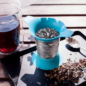 May include: A blue tea infuser with a stainless steel mesh filter and a patterned design. The infuser is sitting on a black tray with loose tea leaves scattered around it.