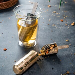 May include: A glass tea infuser with a silver mesh filter filled with loose leaf tea. The infuser is in a clear glass cup with a double wall. The tea infuser is labeled "OLIV." A small silver tea strainer with a handle is filled with loose leaf tea. The strainer is labeled "OLIV."