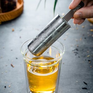 May include: A stainless steel tea infuser with a floral design is being lowered into a glass cup of tea. The infuser has a handle with the word "OLIV" printed on it.