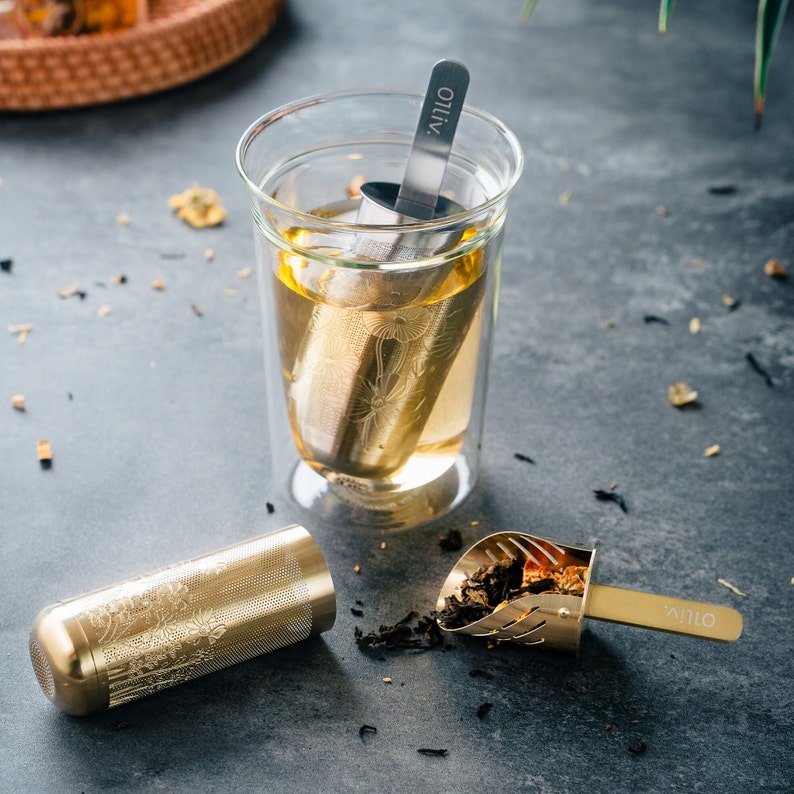 May include: A gold tea infuser with a floral design is submerged in a glass of tea. A matching gold tea scoop with the word "Olivo" on the handle is on the table next to the glass.