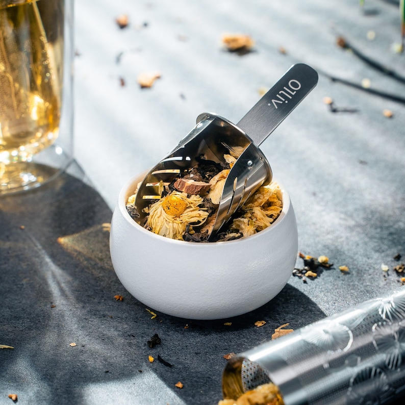 May include: A white ceramic bowl filled with dried flower tea and a silver tea scoop with the word "OLIV'" engraved on the handle.
