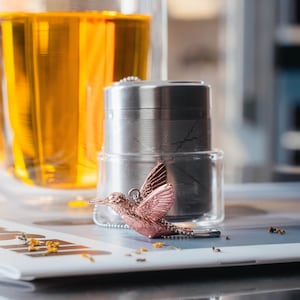 May include: A rose gold hummingbird tea infuser with a stainless steel tea strainer. The infuser is sitting on a white surface with a glass of tea in the background.