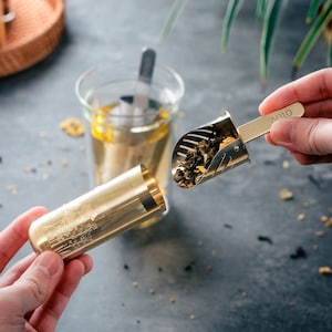 May include: Two gold-colored tea infusers. One is a long, cylindrical shape with a patterned design. The other is a scoop with a handle that says "Oliy". Both infusers are filled with loose leaf tea.