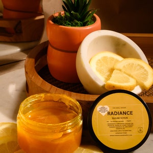 May include: A glass jar of orange sugar scrub with a white label that reads "RADIANCE SUGAR SCRUB" and lists ingredients including cane sugar, coconut oil, argan oil, honey, turmeric, and lemon essential oil. The jar is sitting on a wooden tray with lemon slices and a potted succulent plant.