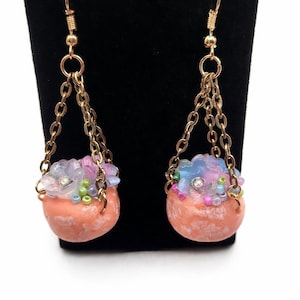 May include: A pair of dangle earrings featuring peach-colored, rounded stone-like pendants. Each earring has a gold-tone chain and hook. The pendants are adorned with clusters of blue, pink, and green beads, and a small clear gem.
