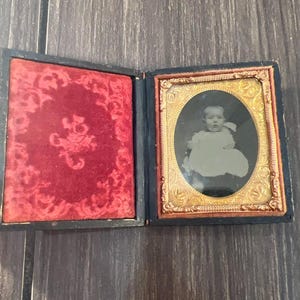 May include: A vintage black and gold framed photograph of a baby in a white dress. The photograph is in a velvet-lined case.