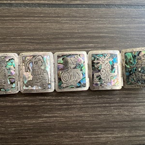 May include: A silver-toned bracelet featuring five rectangular panels. Each panel showcases an engraved figure and is inlaid with iridescent abalone shell. The bracelet has a clasp closure and is likely a piece of jewelry.