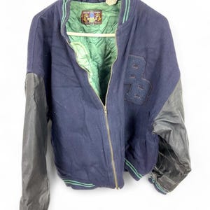May include: Navy blue varsity jacket with black leather sleeves. The jacket features a green and navy striped collar and cuffs, a green quilted lining, and a zipper closure. The letter "PB" is stitched on the front.