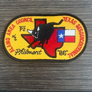 May include: Embroidered patch with a yellow border, featuring a black bull over a red Texas outline. Text includes "Alamo Area Council", "Texas Sesquicentennial", and "Philmont". The Texas flag is also displayed.