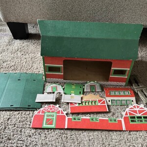 May include: A vintage toy barn set with a red and green color scheme. The set includes a large barn structure, roof panels, and various smaller building components. The barn has green roofing and red walls with white trim. The set is made of cardboard.