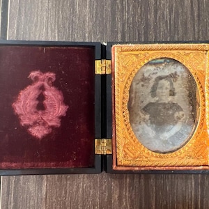 May include: A black wooden frame with a velvet lining and a gold-toned frame with a faded photograph of a person inside.