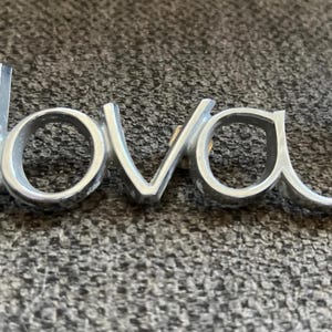 May include: A silver "Nova" emblem with a matching "Chevy" badge. The emblem is in a cursive font and is made of metal. The Chevy badge has a red stripe.
