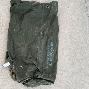 May include: An olive green canvas duffel bag with a drawstring closure and a metal grommet. The bag has a shoulder strap and a printed text that reads "GIBSON R T 243-82-0404". The bag is lying on a gray surface.