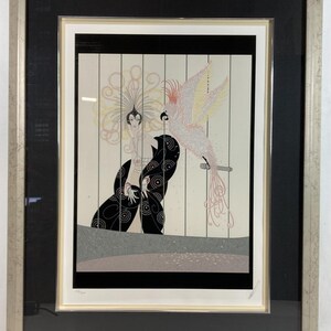May include: A framed print of a woman in a black dress with a large feathered headdress and a pink and white parrot. The woman is standing behind a set of bars, and the parrot is perched on a branch. The background is a light gray with a pattern of small pink flowers.