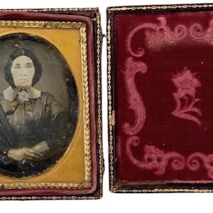 May include: A vintage daguerreotype portrait of a woman in a dark dress with a white collar. The portrait is framed in a gold-colored frame and is set in a red velvet case.