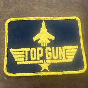 May include: A rectangular patch with a navy blue background and a yellow border. The patch features a yellow jet plane and the words "TOP GUN" in yellow, along with a star. The design is embroidered.