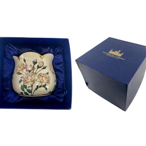 May include: A cream-colored vase with a floral rose design, presented in a blue box with blue satin lining. The vase has a scalloped rim and gold accents. The box is closed and has gold text.