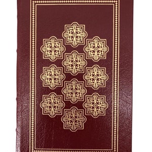 May include: A burgundy-colored book with gold-colored decorative elements. The cover features a gold border and a pattern of nine gold floral designs. The book's spine is visible on the left side.